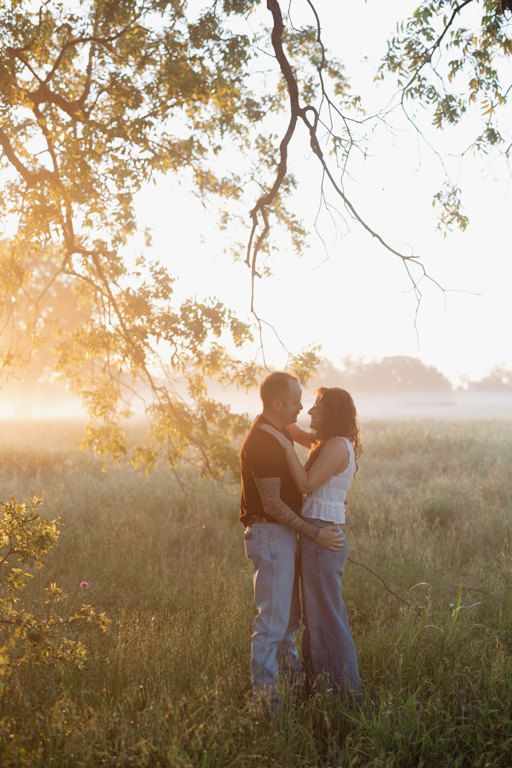 Couple embracing during a dreamy sunrise engagement session in Waco, Texas surrounded by tall grass and morning fog